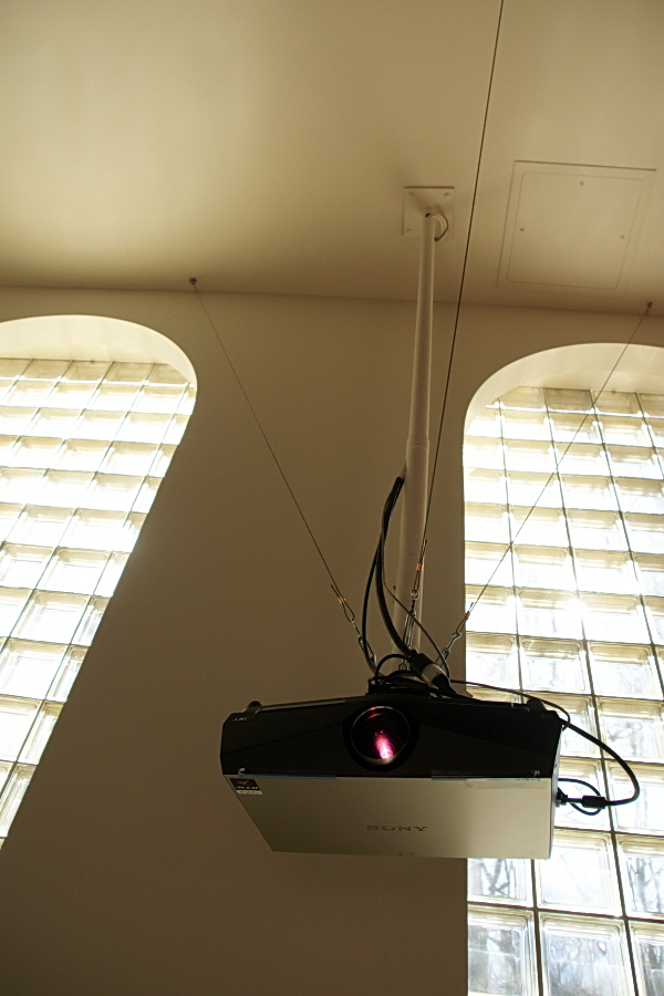 A projector is mounted to a long pole extending from the ceiling. Three stabilizing wires extend from where the projector attaches to the pole up to the ceiling, forming a triangle around the pole