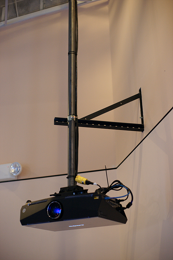 A projector is mounted to a long pole attached to the ceiling. A metal brace extends from the wall and attaches to the pole to stabilize it