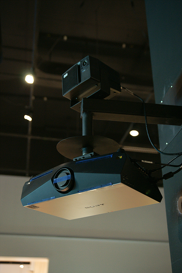 A projector is mounted a short pole that extends down from a square metal arm extending out from the wall. A speaker is mounted above the projector
