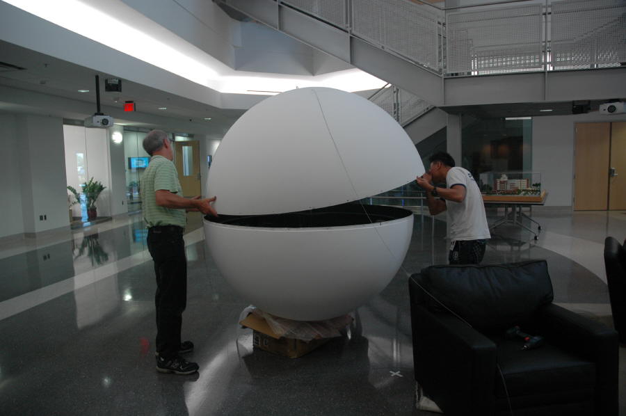 Two people are seen placing the top half of a Science On a Sphere onto the bottom half